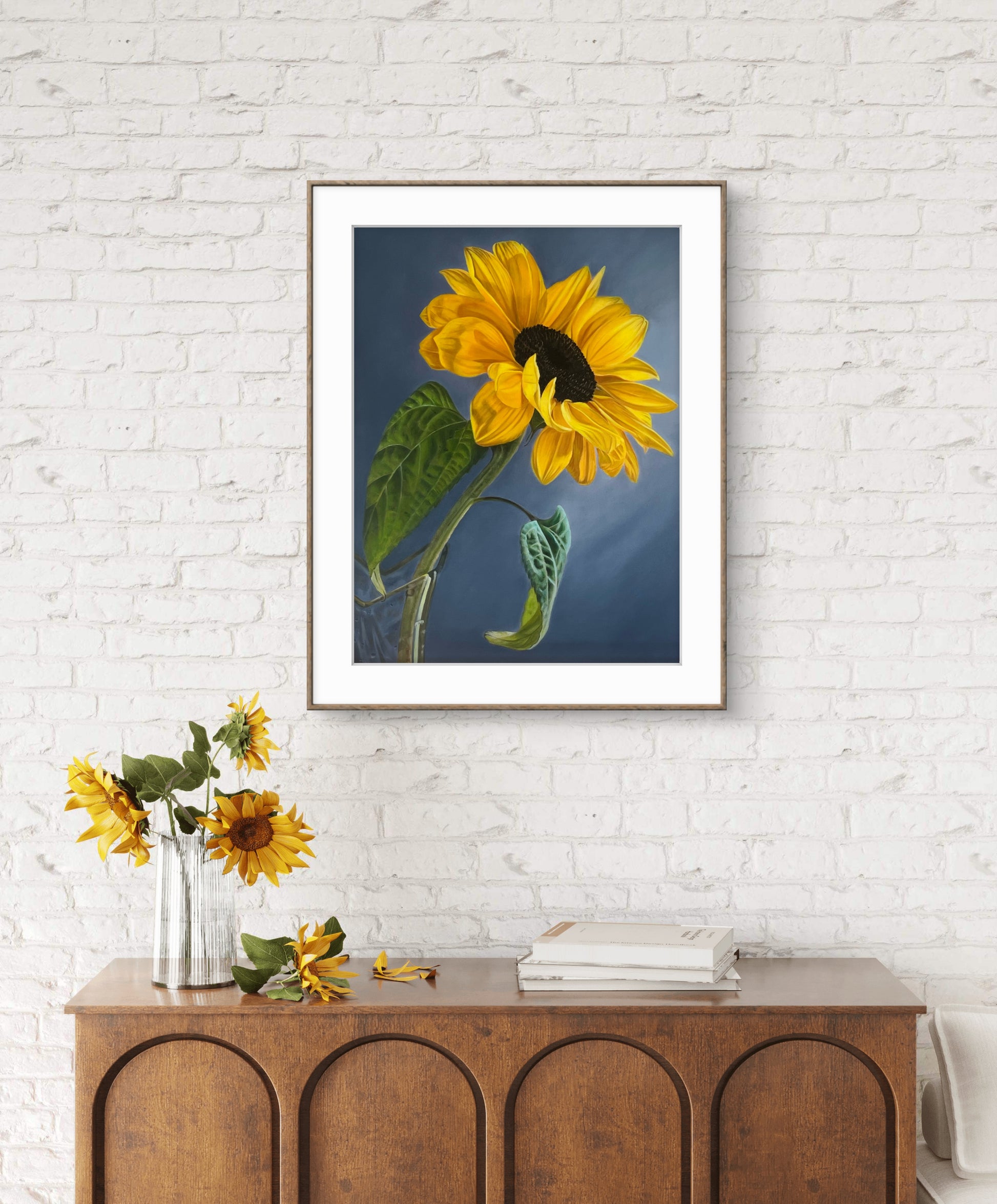 Framed picture of a sunflower on a white brick wall with a wooden cabinet below.